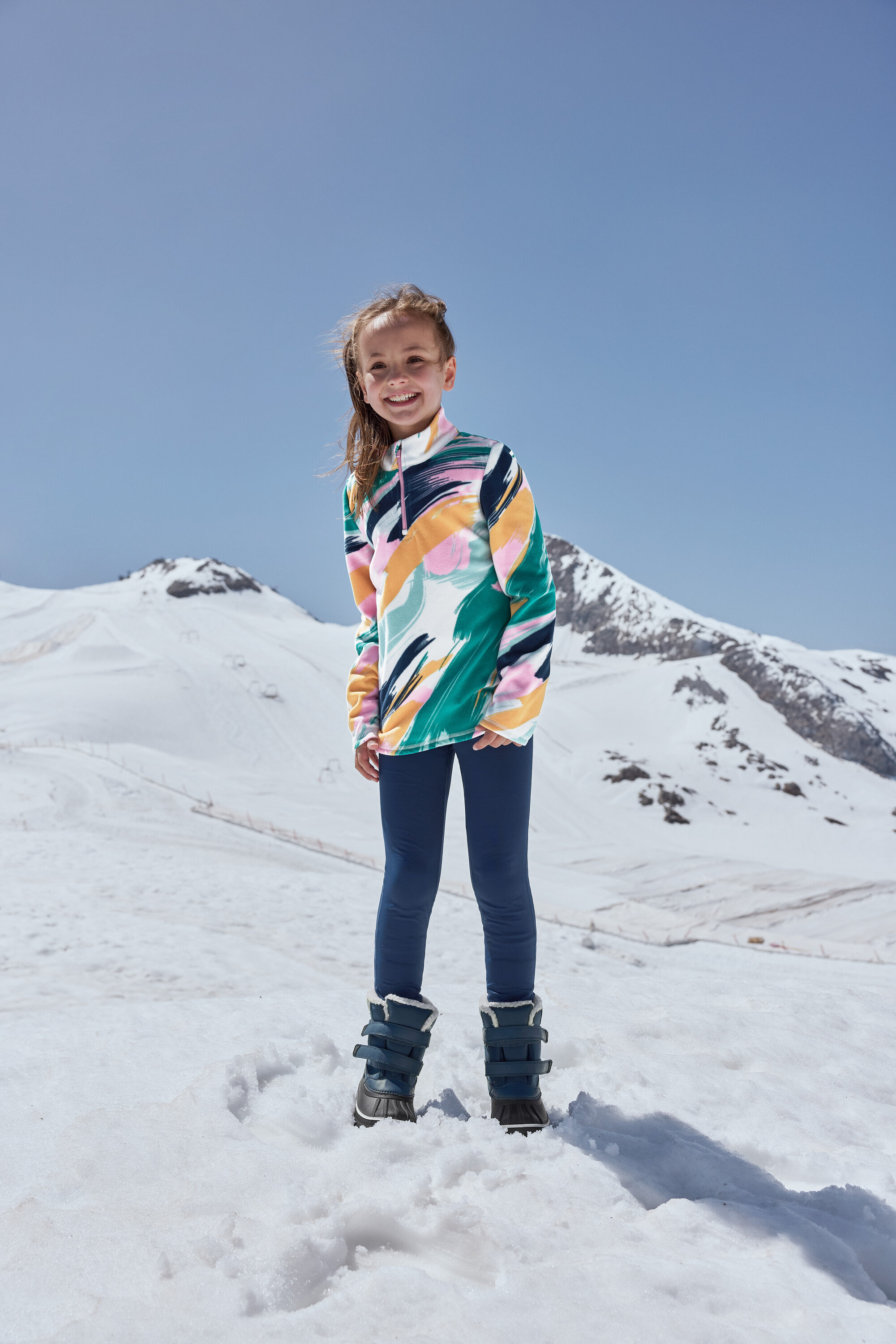 Egy mosolygós lány áll a hóban, kék, minden időjáráshoz való gyermekcsizmát visel.