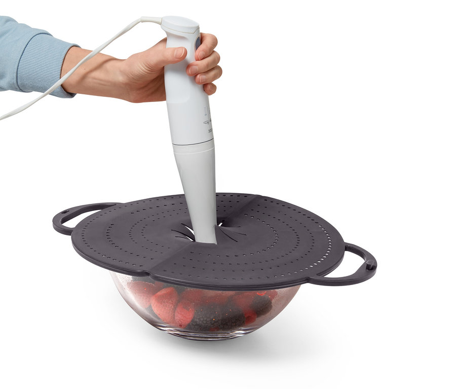 Egy kéz egy fehér botmixert tart egy gyümölccsel teli tál fölé, amelyet egy lyukacsos szürke fedő fed le.