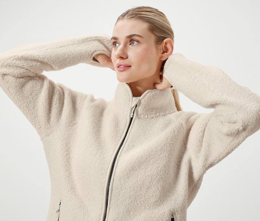 Egy szőke hajú, lófarokba kötött nő pózol egy krémszínű, cipzáras polár pulóverben.