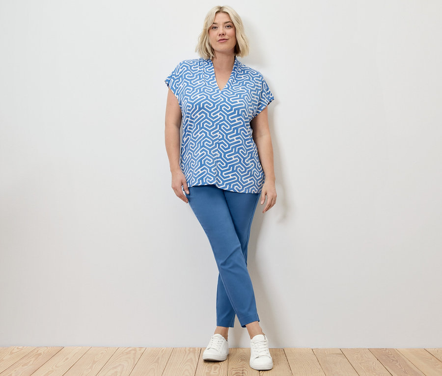 Egy szőke nő pózol kék sztreccsnadrágban és kék V-nyakkivágású blúzban.
