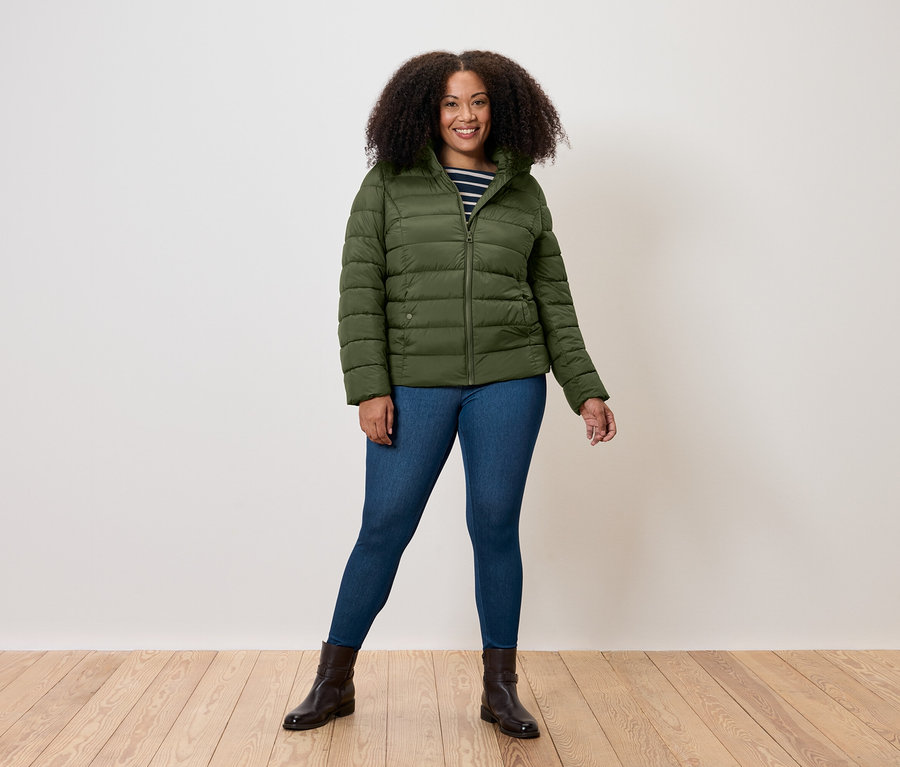 Egy nő áll olívazöld színű steppelt kabátban, farmerben és barna csizmában.