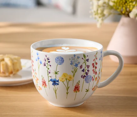 Virágos bögre kávéval és latte art szívvel egy faasztalon, egy tányér sütemény és egy váza virág mellett.