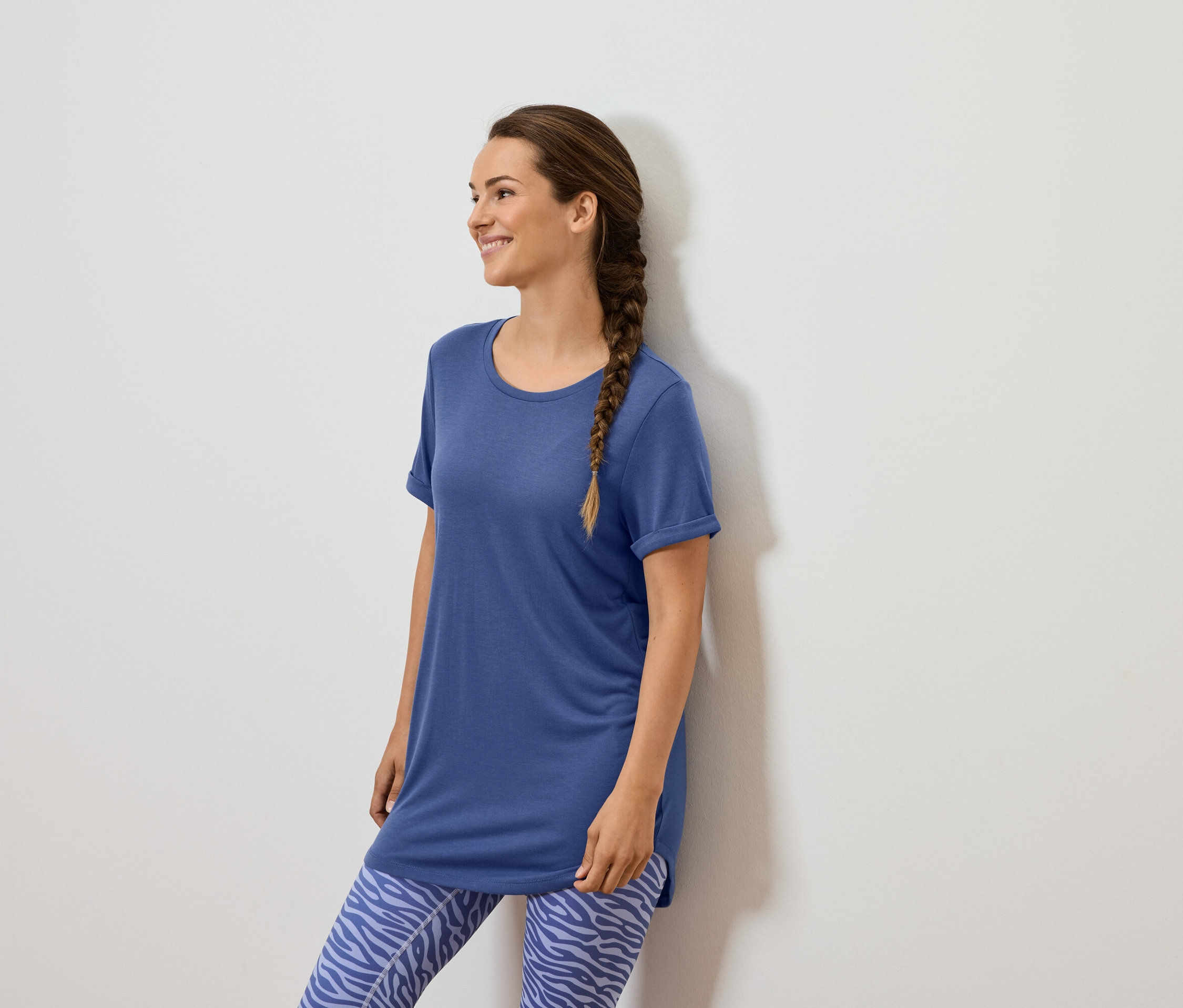 Egy barna copfos nő áll egy fehér fal mellett, kék pólót és kék, állatmintás leggingset visel.