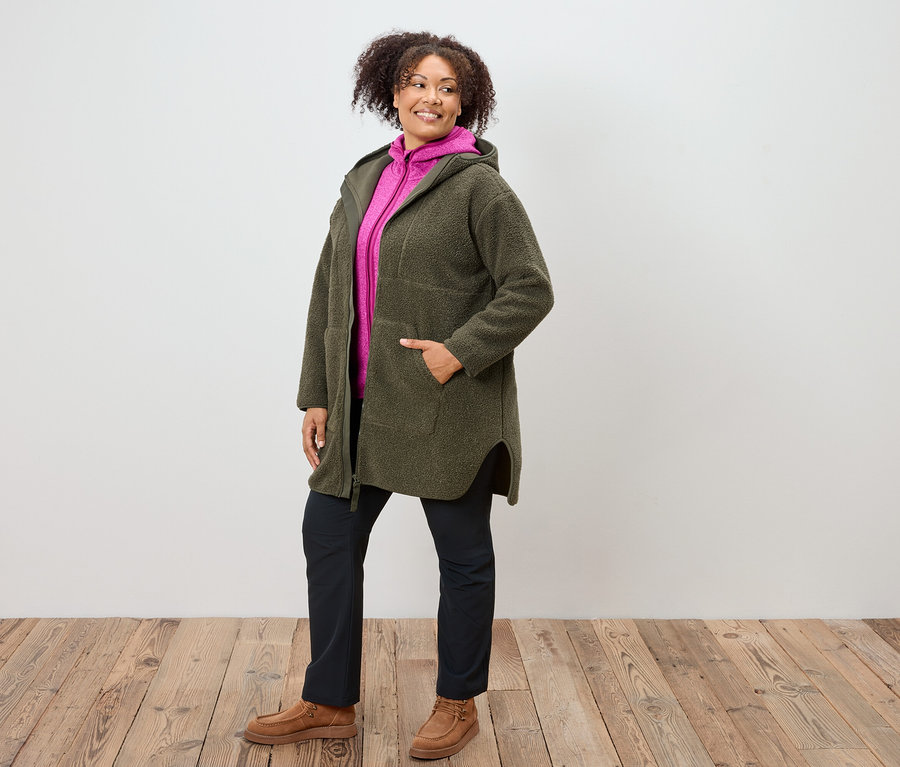 Nő olívazöld kapucnis kabátban, rózsaszín pulóverben, sötétkék nadrágban és barna cipőben áll fa padlón.