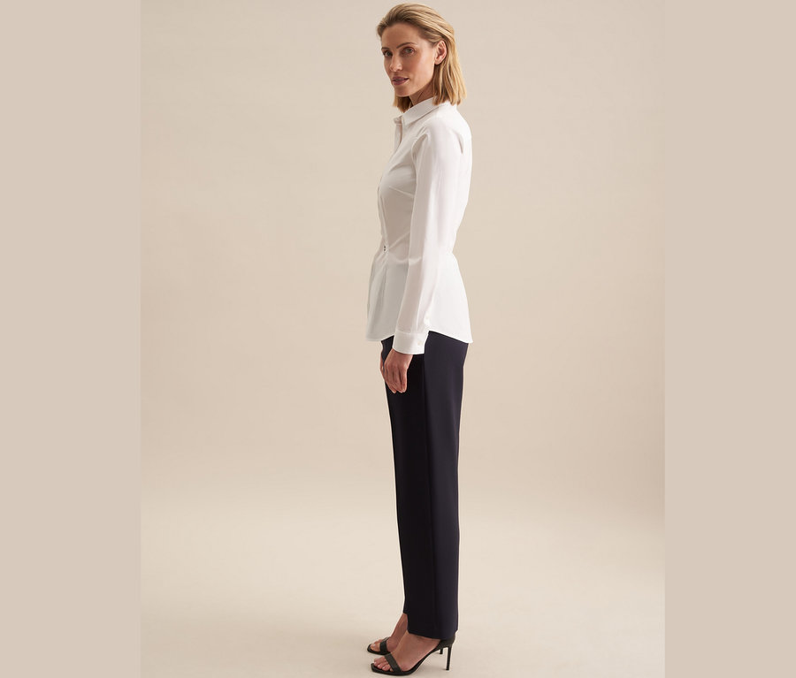 Nő áll oldalt, fehér Seidensticker slimfit blúzt és sötétkék nadrágot visel.