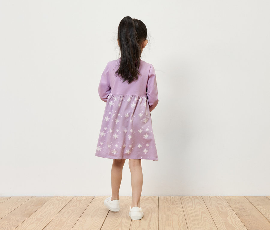 Egy kislány lila, csillagos ruhában és fehér tornacipőben áll háttal a kamerának.