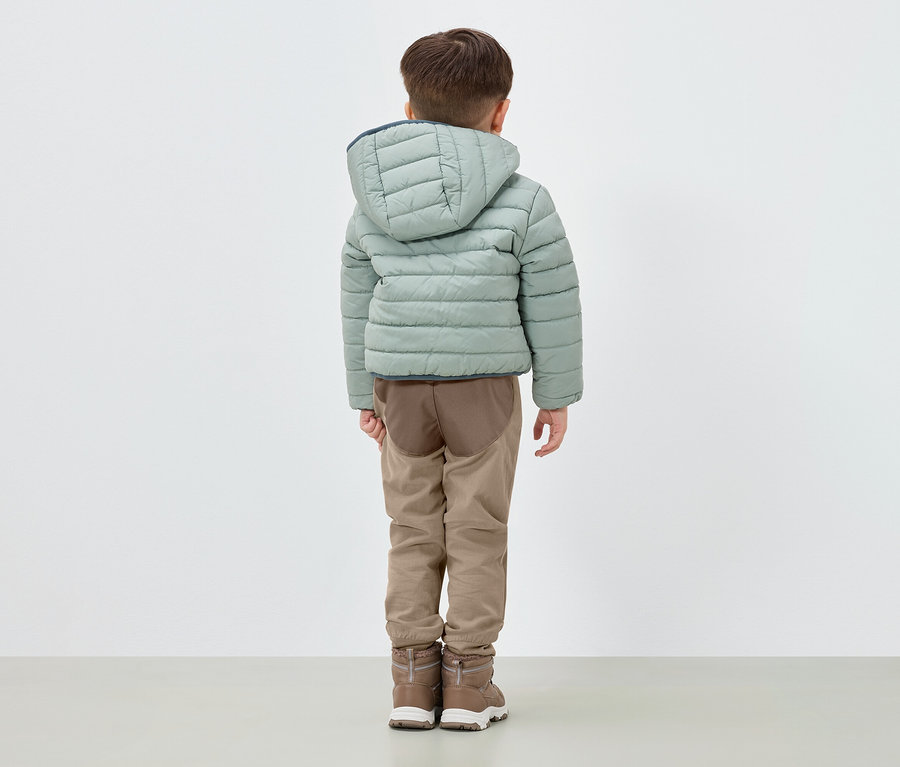 Egy fiú háttal áll, világoszöld, kapucnis pehelykabátot, barna nadrágot és barna csizmát visel.