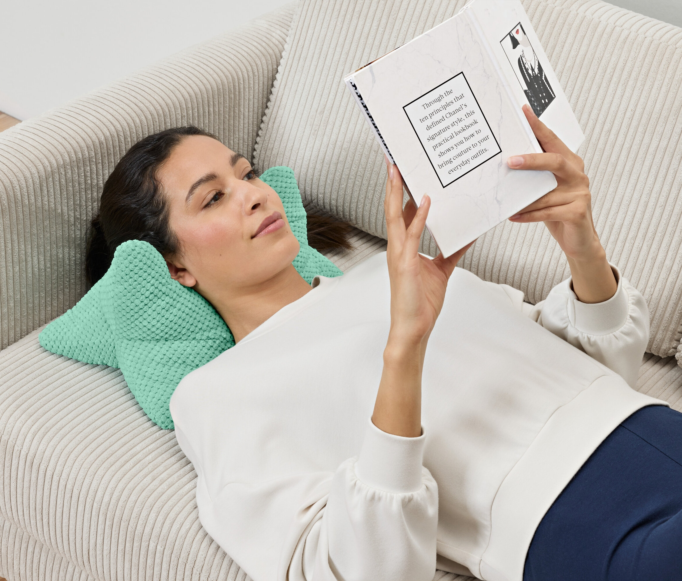 Egy nő a kanapén fekszik és könyvet olvas, a feje egy olvasópárnán pihen.