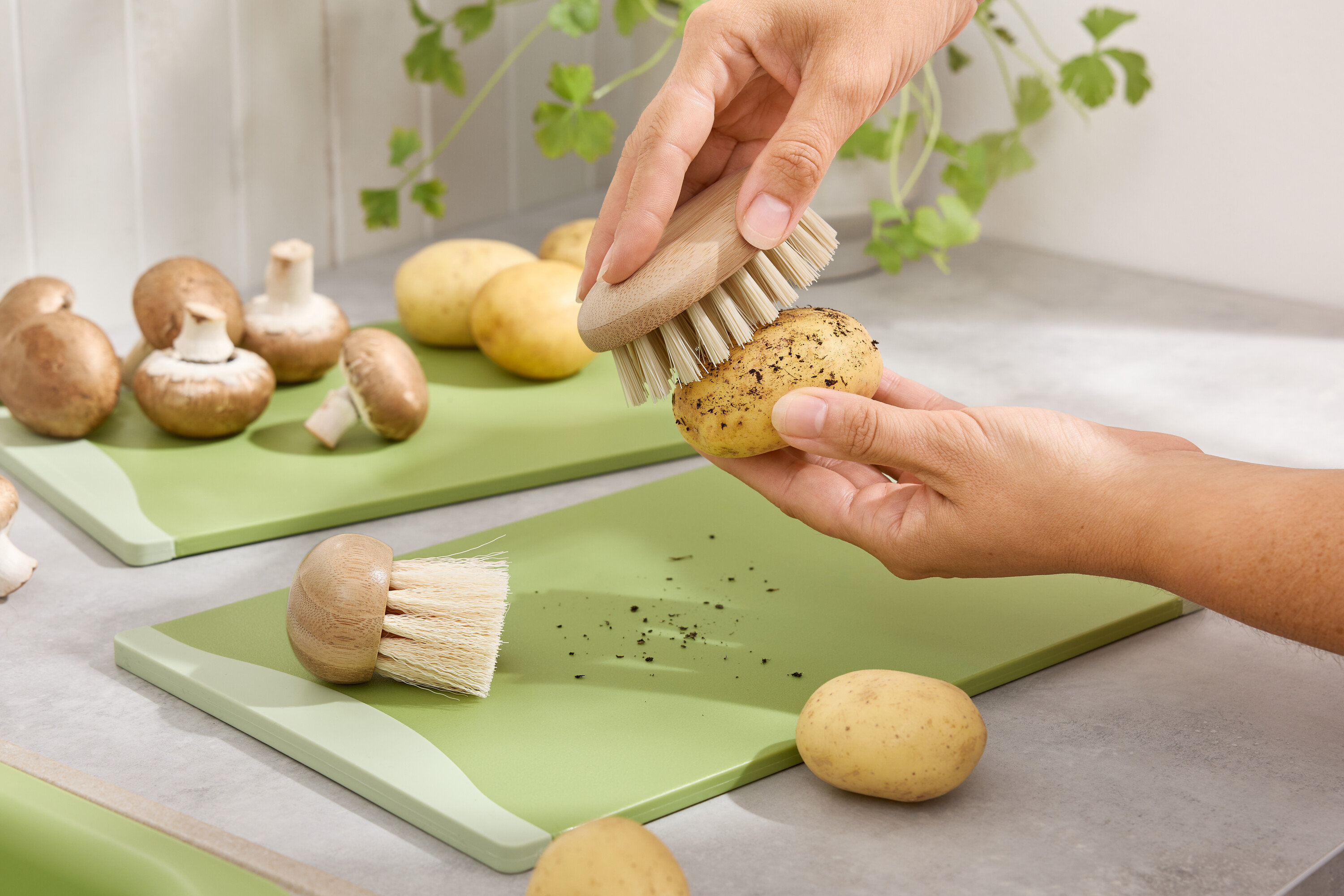 Kezek zöld vágódeszkán krumplit tisztítanak kefével. Barna csiperkegomba és krumplik a háttérben.