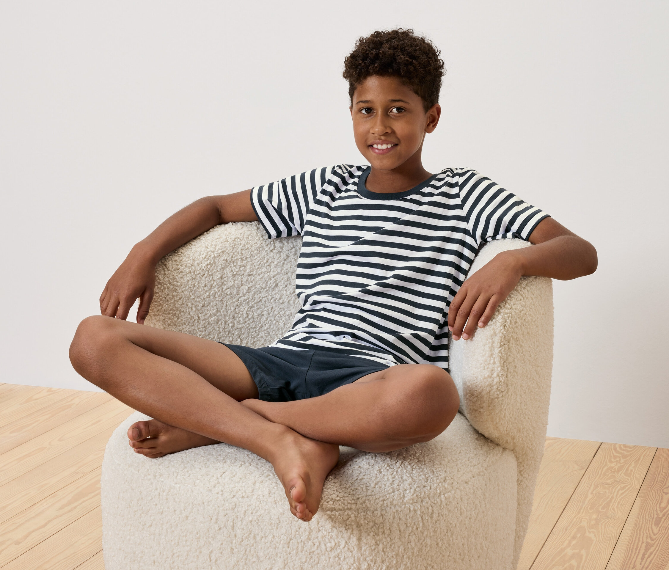 Egy fiatal fiú keresztbe tett lábakkal ül egy krémszínű fotelben. Csíkos pólót és sötétkék rövidnadrágot visel.