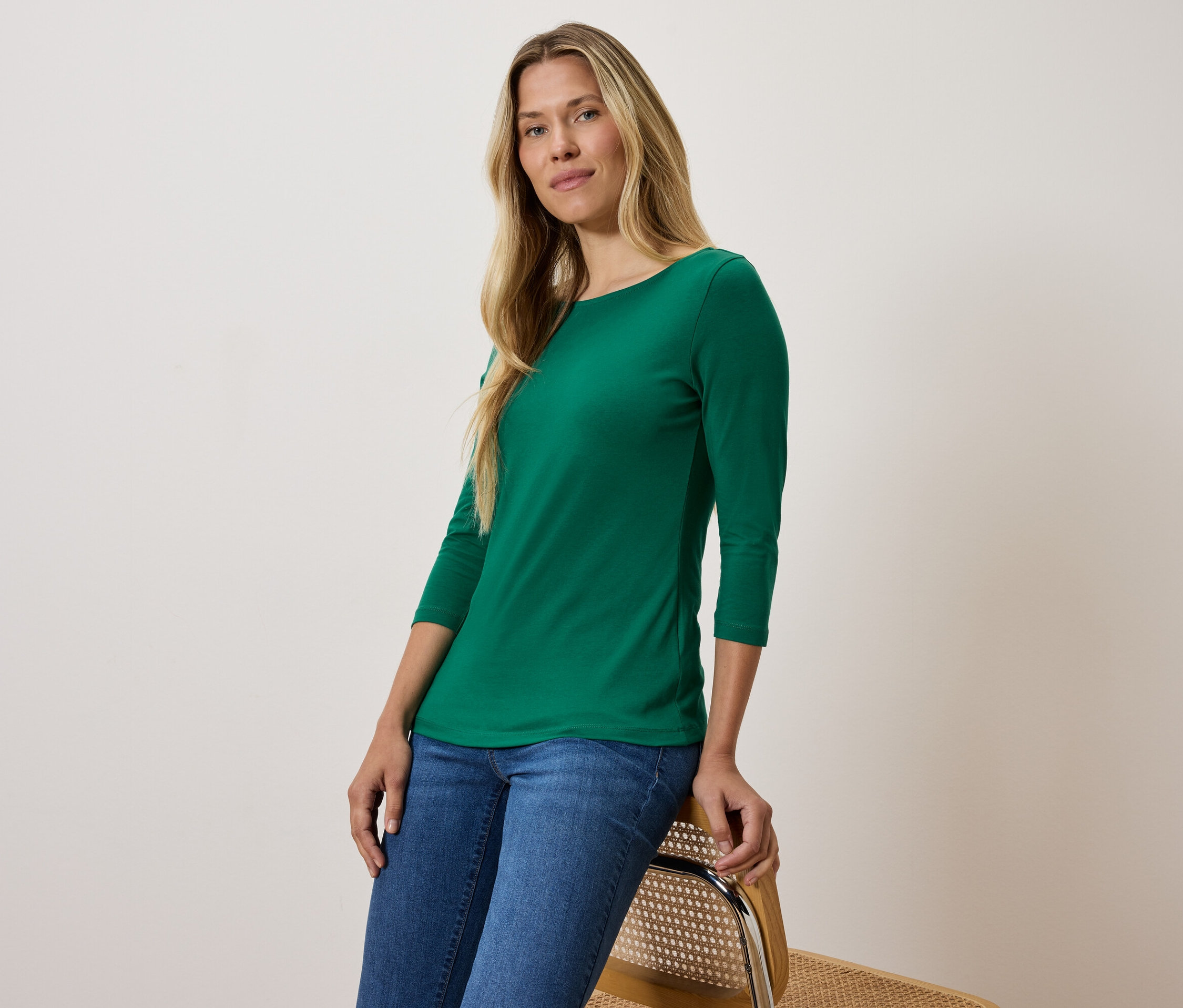 Szőke nő zöld háromnegyedes ujjú pólóban és kék farmerben ül egy széken.