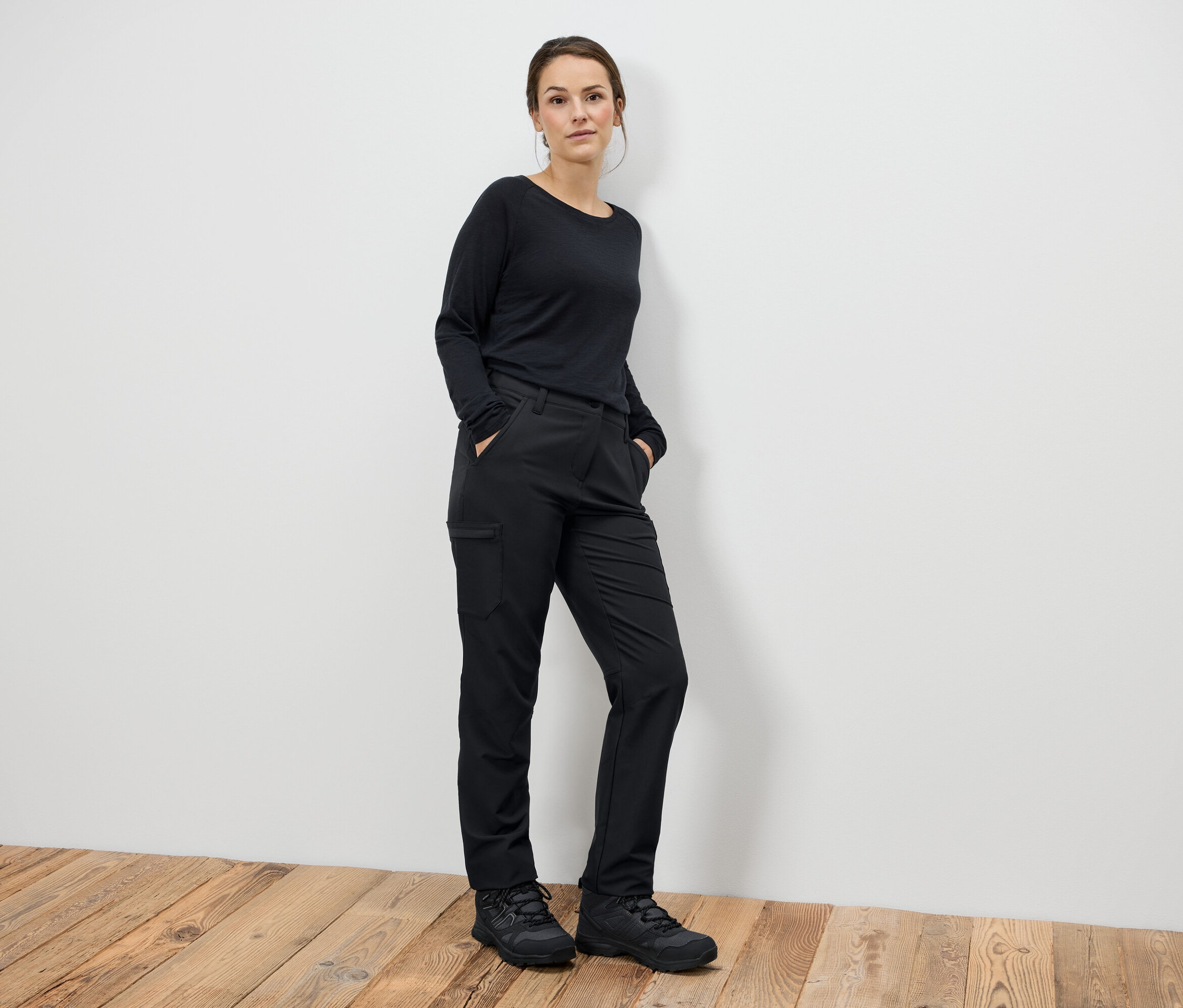 Egy nő áll fekete nadrágban és fekete csizmában, kezét a zsebében tartva.