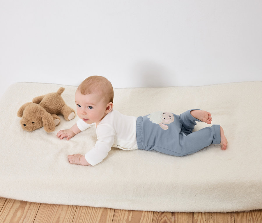 Egy baba fekszik a hasán egy fehér alátéten, két barna plüsskutya mellett.