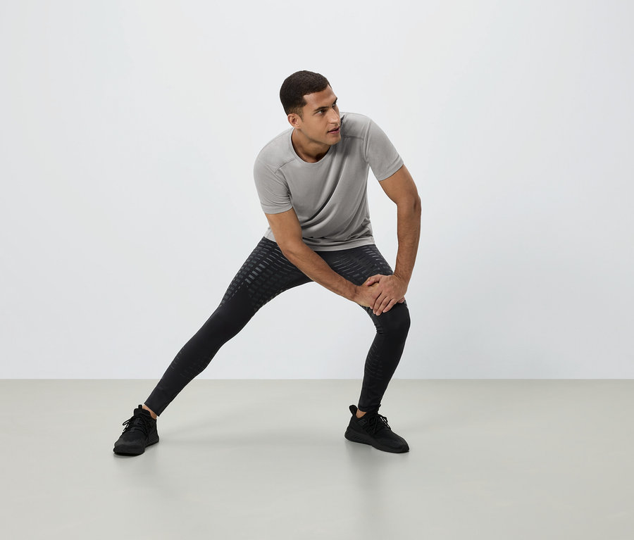 Egy férfi nyújtózkodik szürke pólóban, fekete leggingsben és fekete tornacipőben fehér háttér előtt.