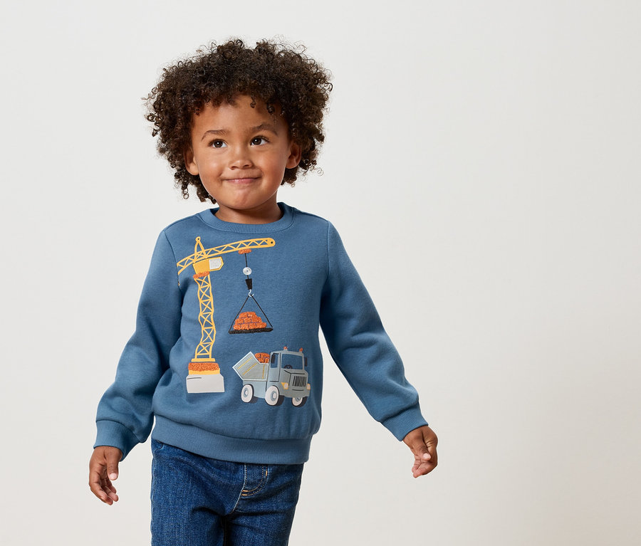 Egy göndör hajú fiú kék pulóvert visel, rajta egy sárga daru és teherautó applikációval.