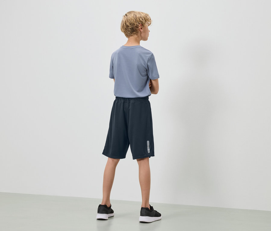 Egy szőke, göndör hajú tinédzser áll háttal a kamerának, kék pólót, sötétkék rövidnadrágot és fekete tornacipőt visel.