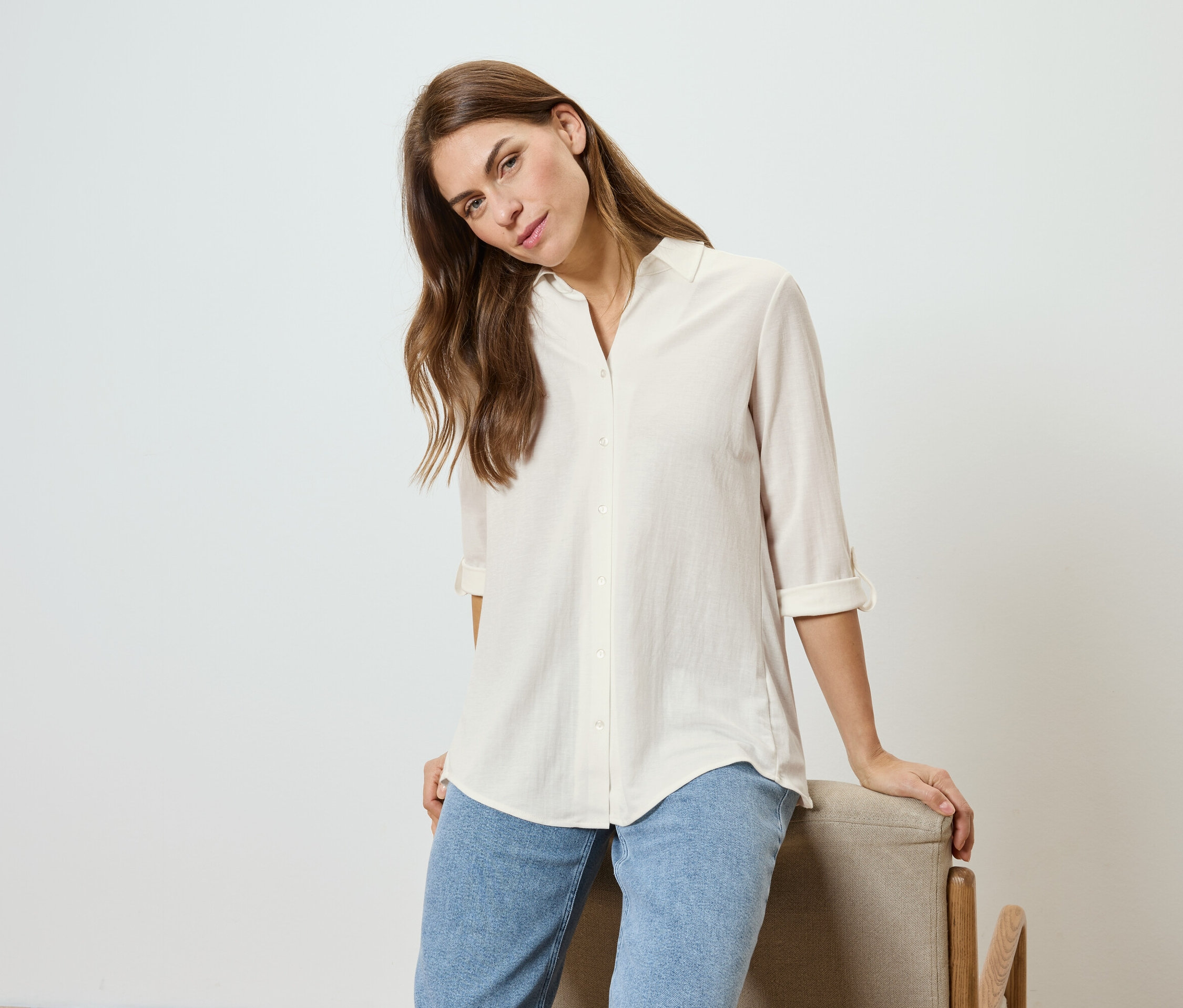 Hullámos hajú nő világos inget és farmert visel, egy széknek támaszkodik.