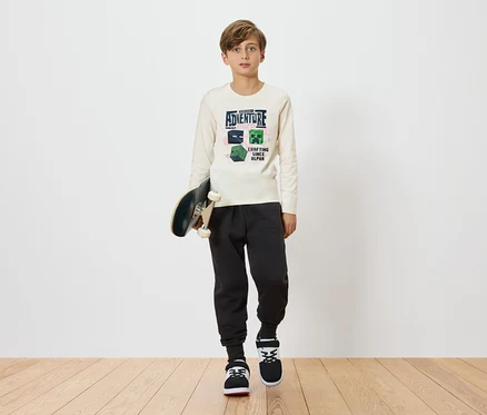 Egy fiú gördeszkát tart, világos, Minecraft mintás hosszú ujjú pólót, sötét színű jogging nadrágot és tornacipőt visel, egyszerű háttér előtt.