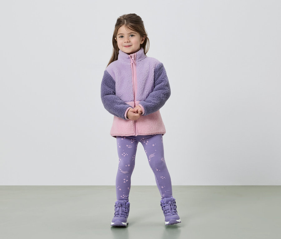 Egy kislány áll összekulcsolt kézzel, lila és rózsaszín polárkabátot, csillagos leggingset és lila csizmát visel.