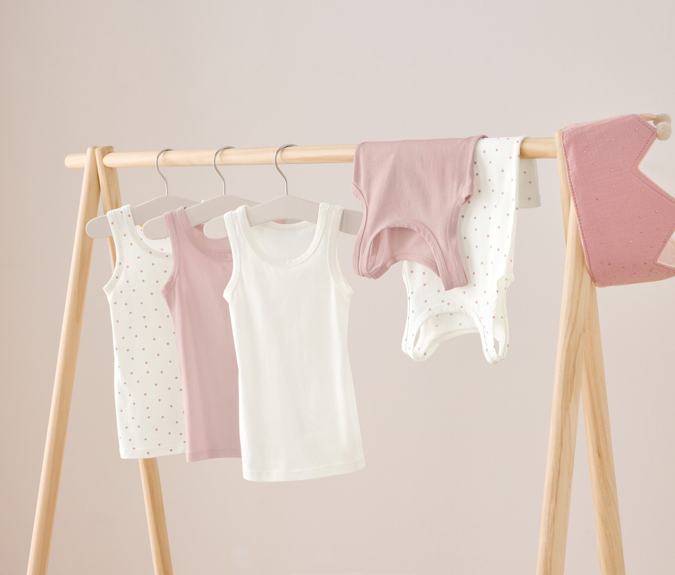 Egy fa állványon három trikó lóg fehér, rózsaszín és fehér pöttyös színben, valamint egy rózsaszín ing és egy fehér pöttyös body. Jobb oldalon egy rózsaszín korona lóg.