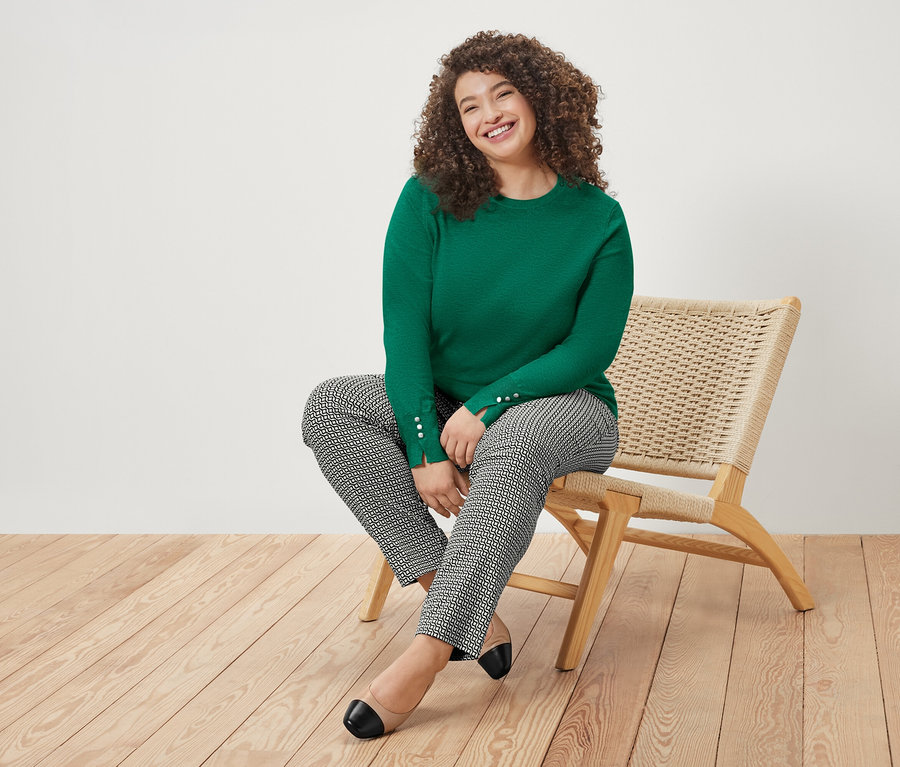 Göndör hajú nő ül egy széken, zöld pulóvert és mintás nadrágot visel.