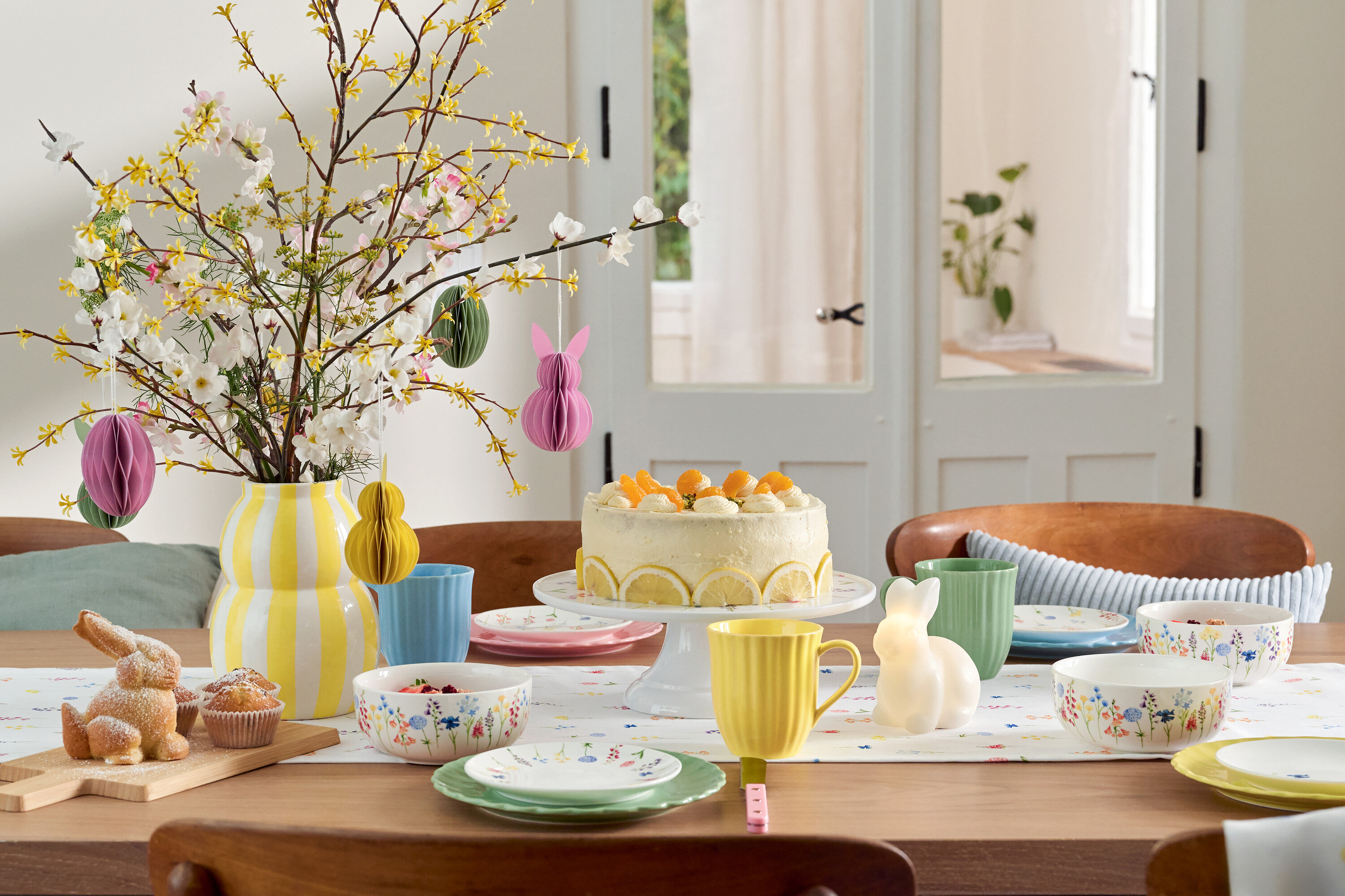 Étkezőasztal húsvéti díszekkel, süteménnyel, edényekkel és virágvázával.