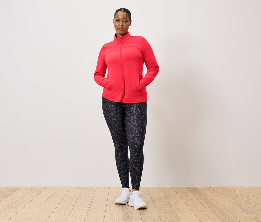 Nő piros kabátban és mintás leggingsben pózol egy fal előtt.