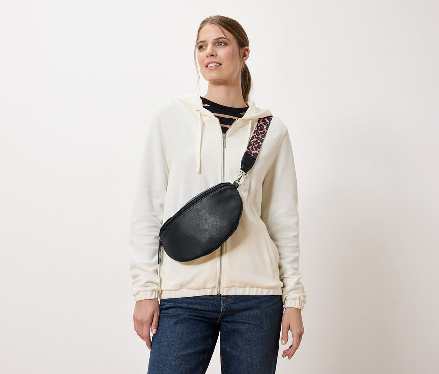 Egy nő fekete övtáskát visel leopárdmintás pánttal, krémszínű kabáton és farmernadrágon átvetve.