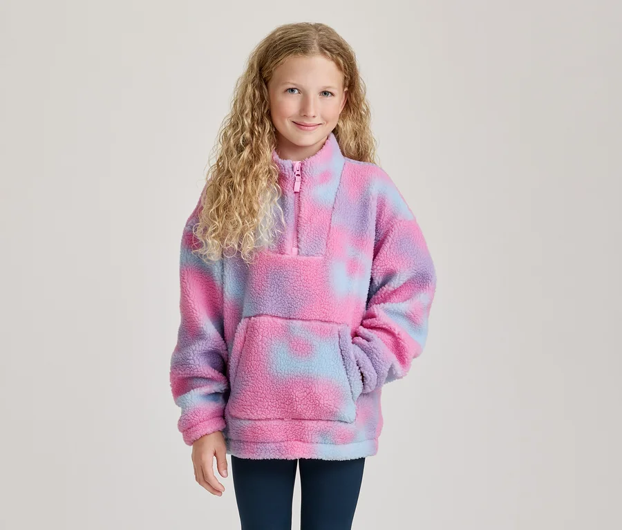 Mosolygó, göndör szőke hajú lány rózsaszín-lila-kék tie-dye polár pulóvert visel félcipzárral és elülső zsebbel.