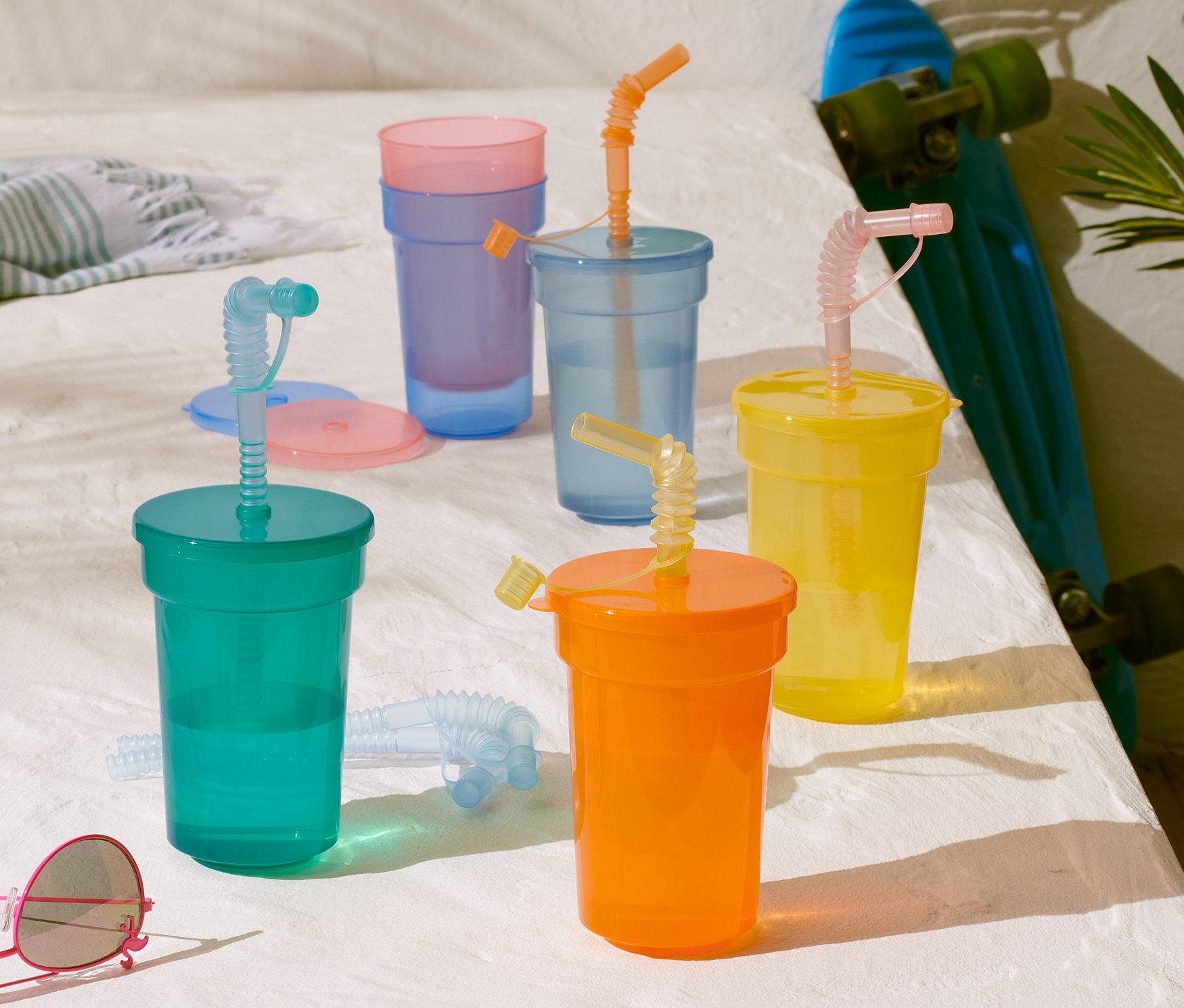 Színes gyermekpohár szett szívószálakkal egy fehér asztalon, egy gördeszka és egy napszemüveg mellett.