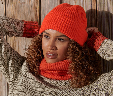 Göndör hajú nő pózol, kezeit a feje mögött tartva, narancssárga kötött sapkát visel.