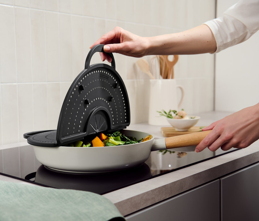 Egy kéz egy 4 az 1-ben konyhai mindenes eszközt tart egy zöldséges serpenyő fölött.