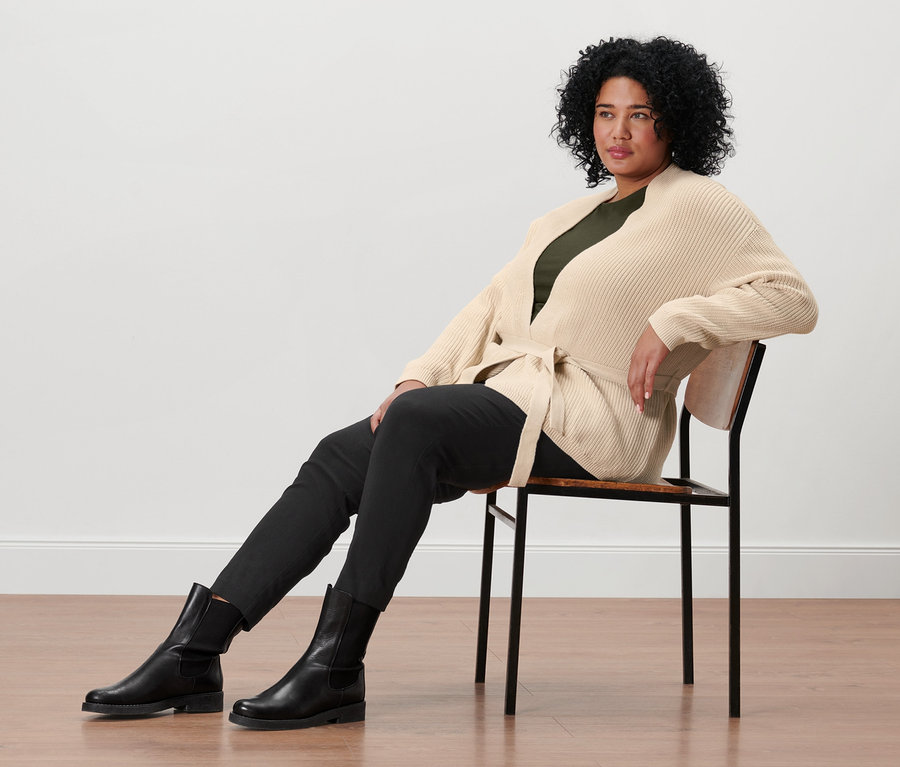 Egy nő ül egy széken, és átlapolós kardigánt, olívazöld, 3/4-es ujjú felsőt, fekete sztreccsnadrágot és bőrcsizmát visel.