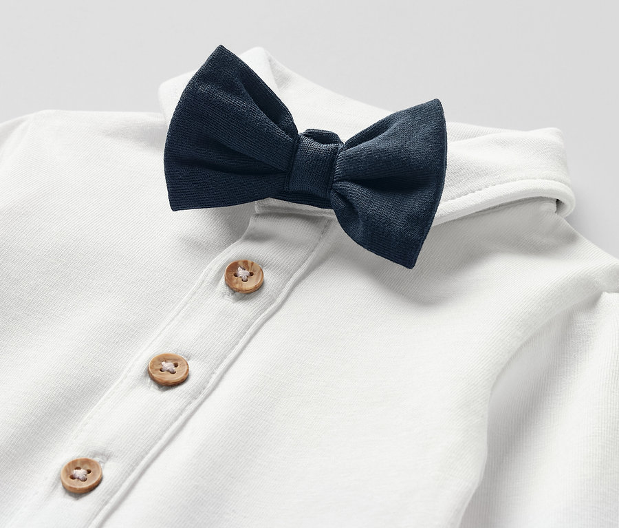 Közeli kép egy fehér, csokornyakkendős kisgyerek bodyról, fa gombokkal.