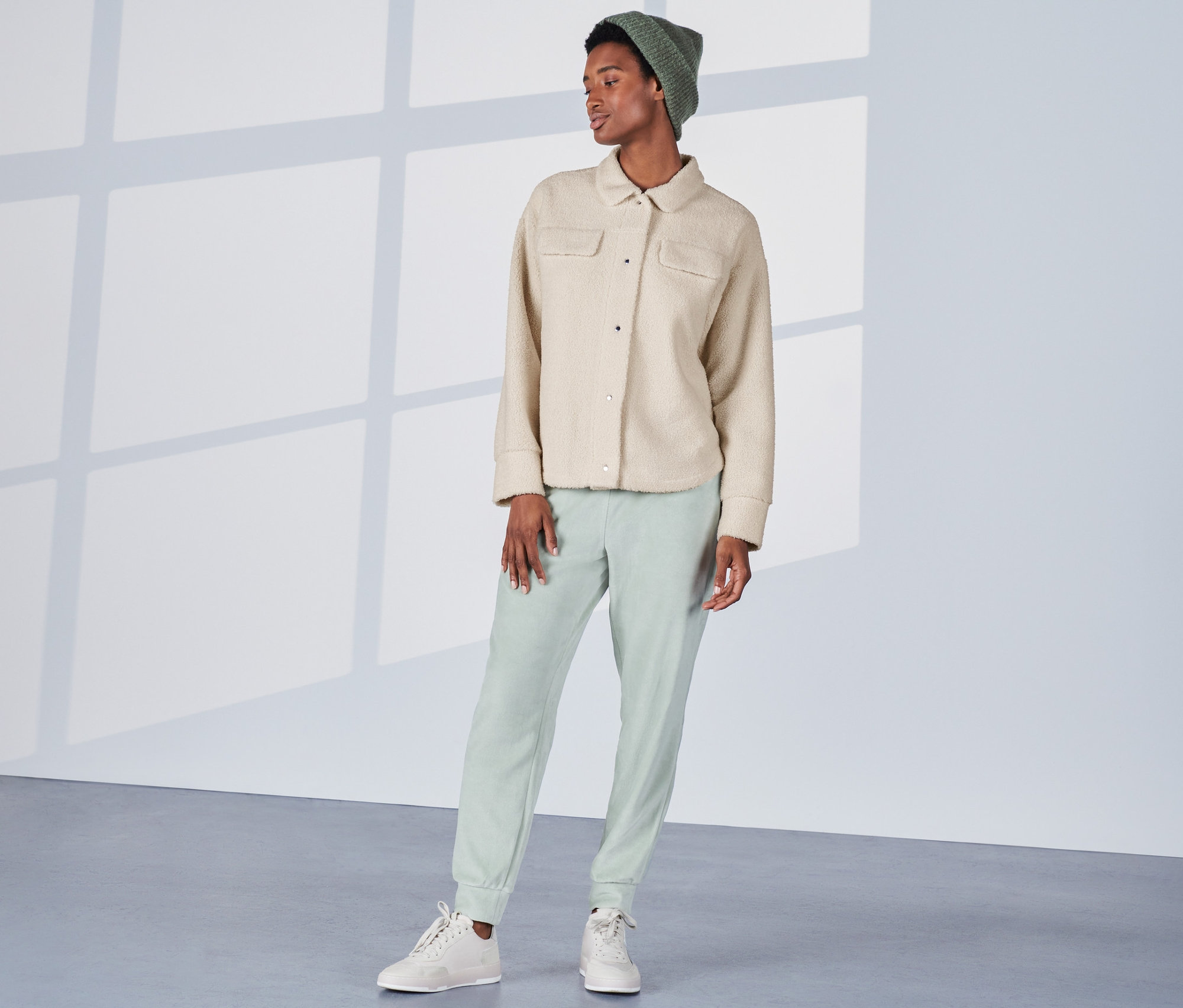 Nő áll, krémfehér plüss ingkabátot, zöld nadrágot és sapkát visel.