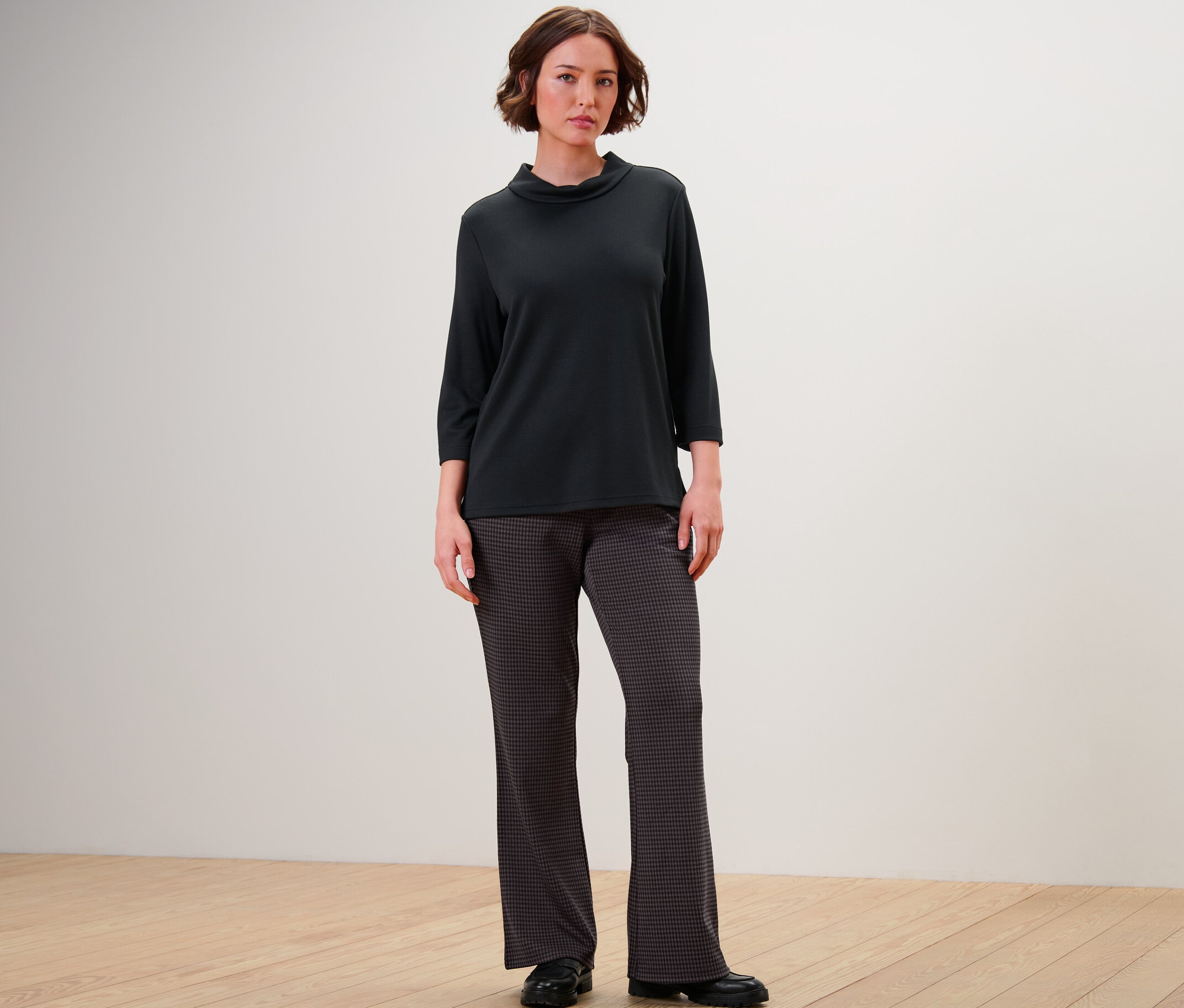 Egy nő áll egy fekete háromnegyedes ujjú felsőben és egy női trapéz nadrágban, tyúklábmintás.