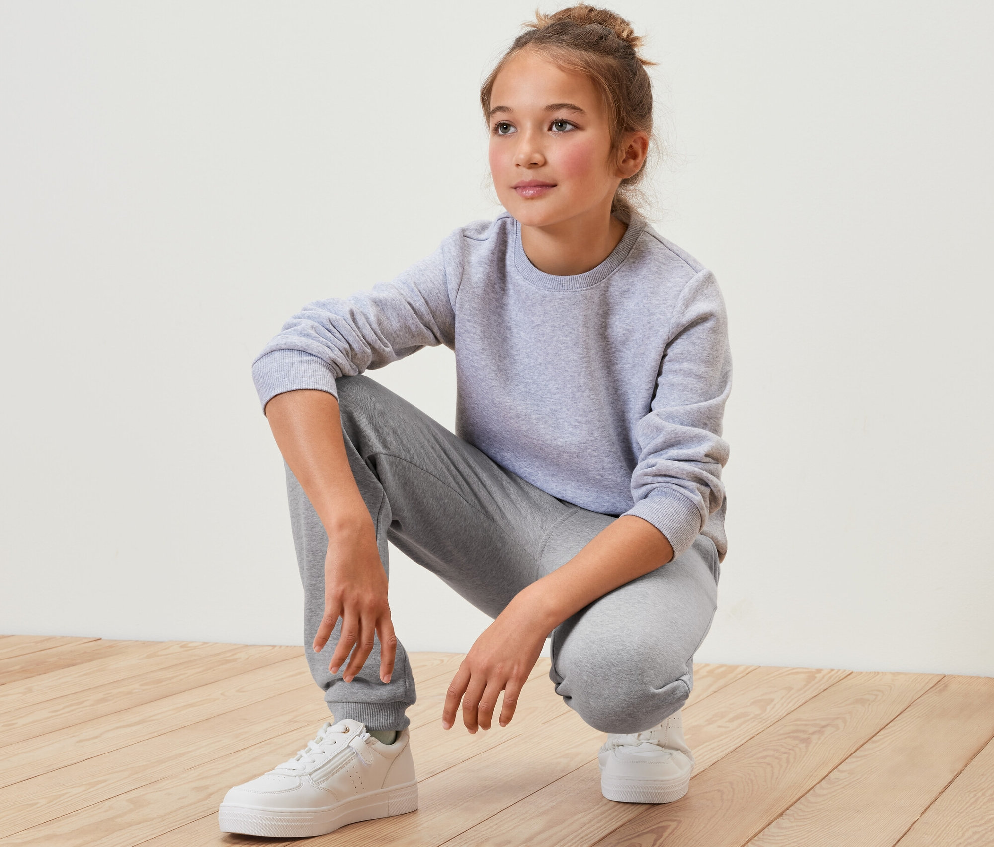 Egy szürke melegítőben és fehér cipőben guggoló lány egy fapadlón. Gyerek szabadidőnadrág van rajta, szürke.