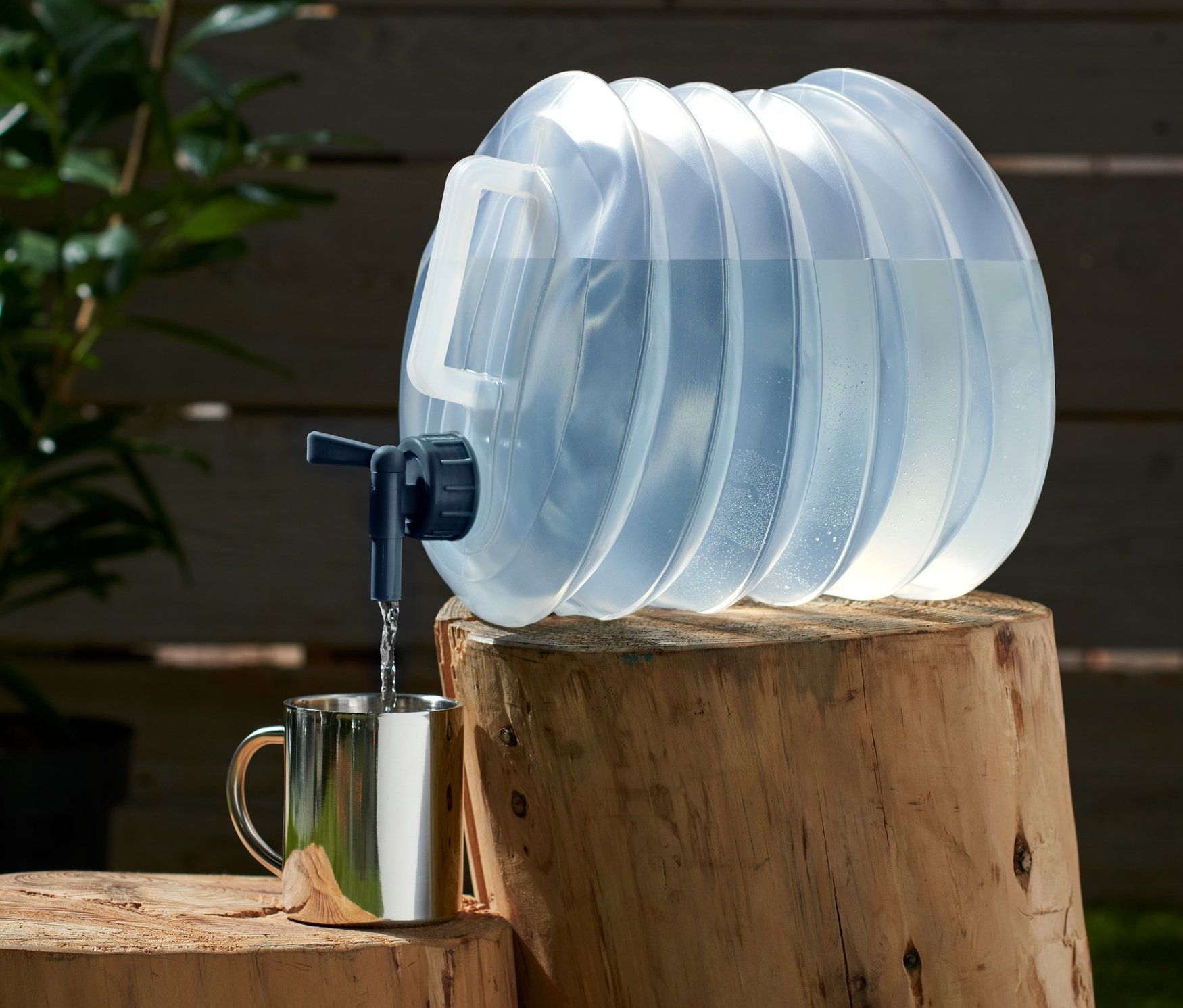 Egy kb. 10 literes, összecsukható vizeskanna egy fatuskón áll, és vizet tölt egy ezüst színű bögrébe.