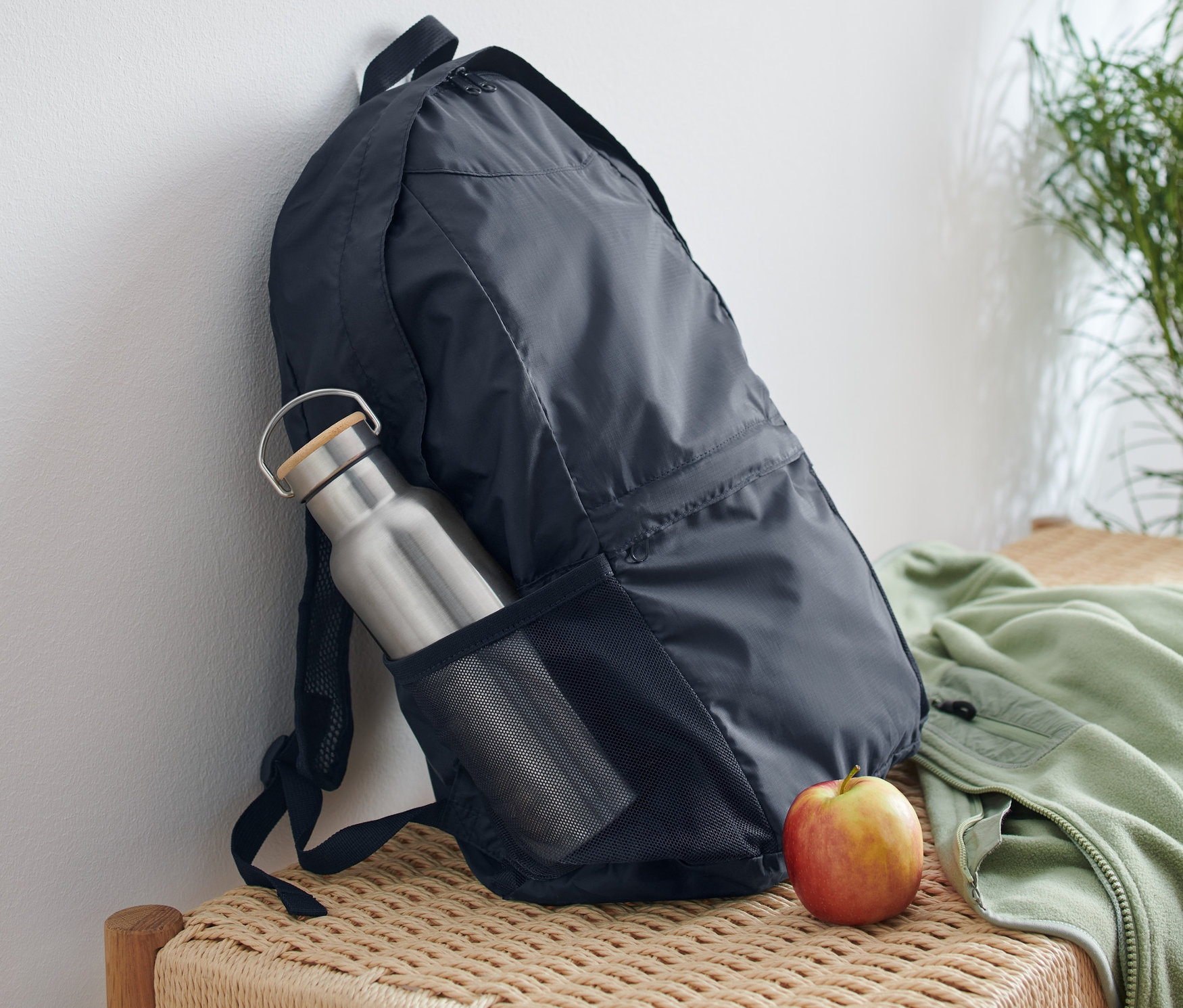 Egy összehajtható hátizsák egy fehér falnak van támasztva, egy vizes palackkal az oldal zsebében és egy almával mellette egy fonott padon.