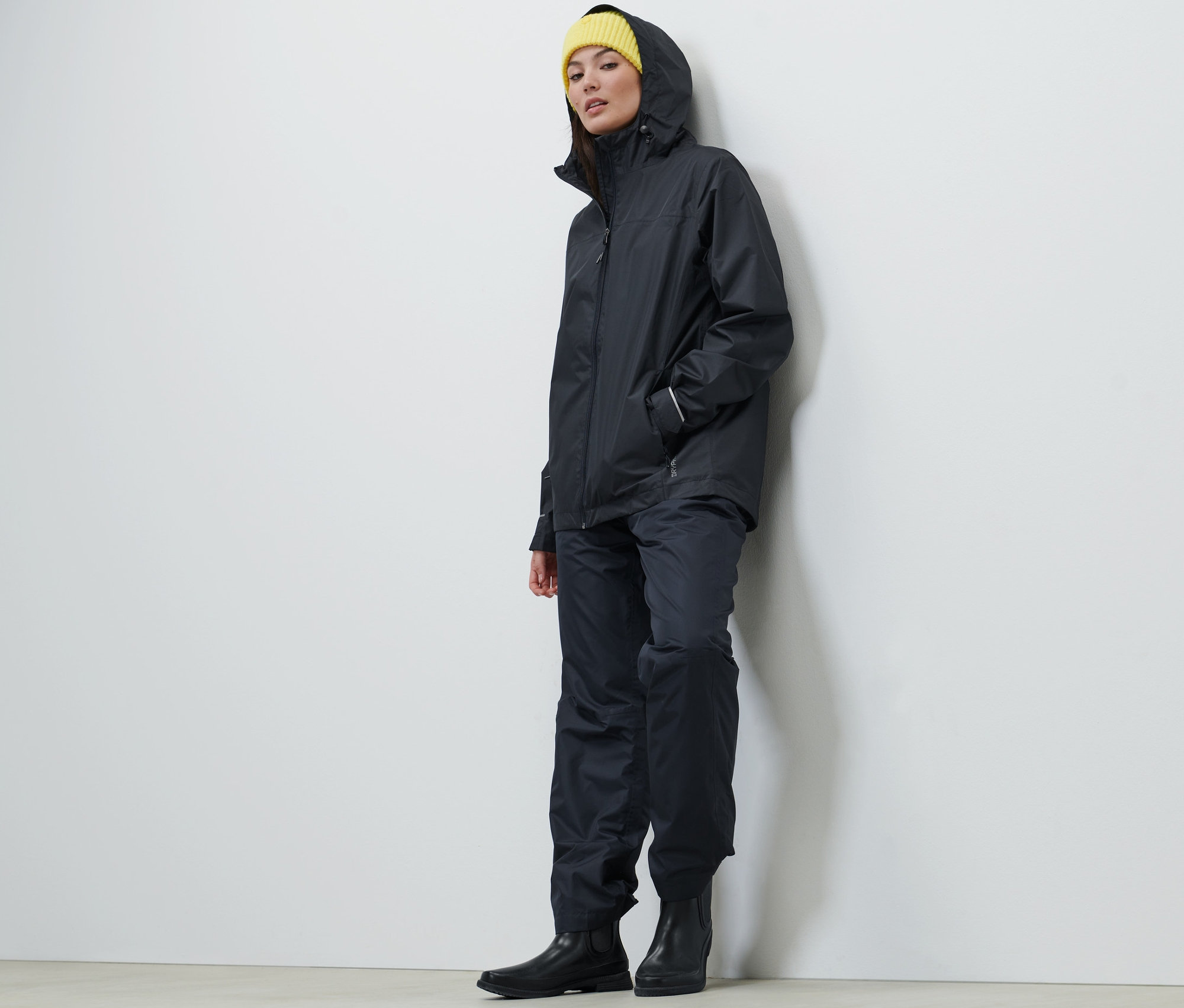 Nő visel sötétkék unisex esőkabátot, nadrágot és sárga sapkát.