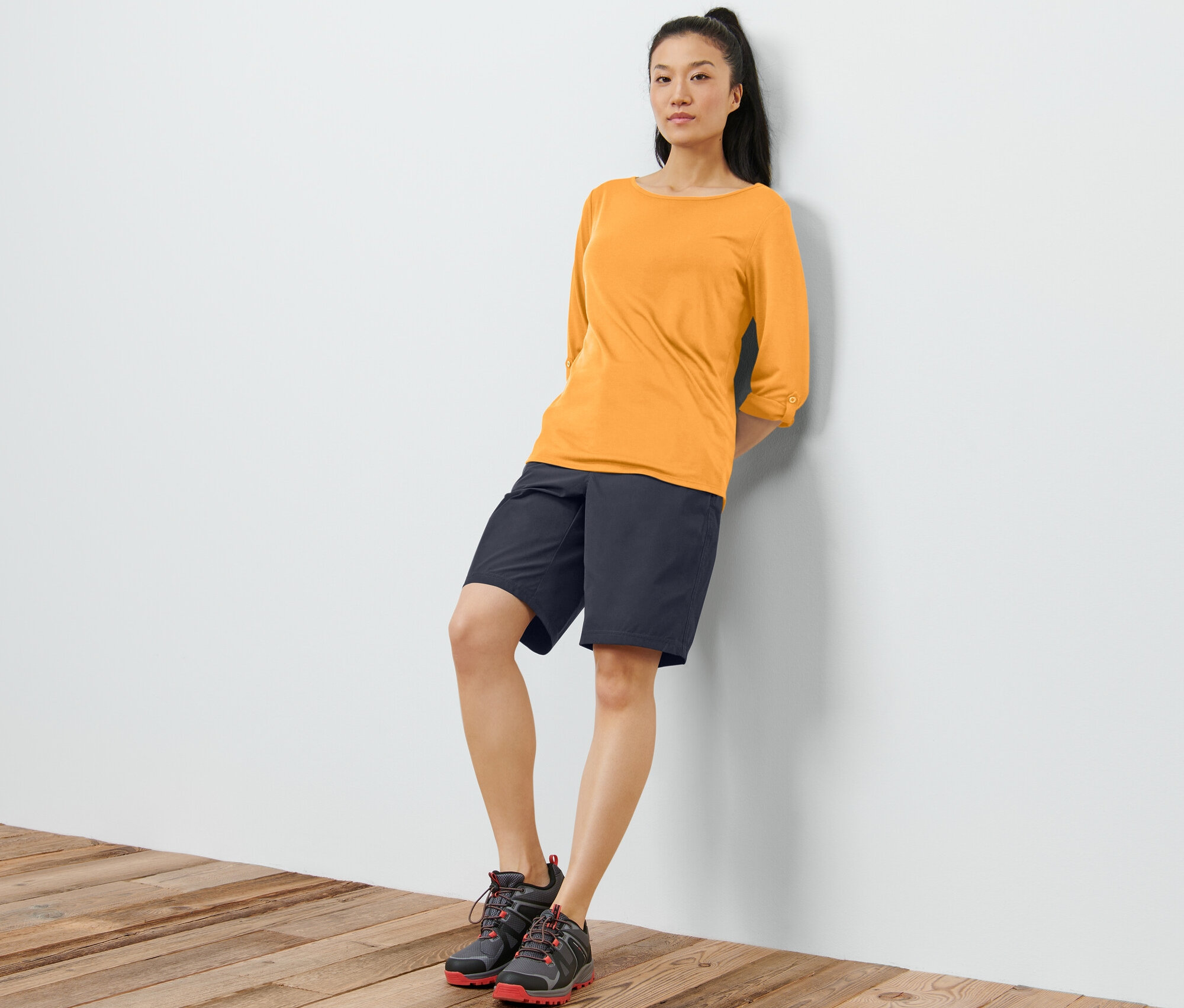 Egy falnak támaszkodó nő narancssárga, 3/4-es ujjú funkcionális felsőt, fekete rövidnadrágot és cipőt visel.