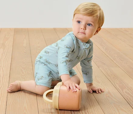 Egy csecsemő fiú mászik a fapadlón, kék, 80-as UV-szűrővel ellátott, sárkányos kezeslábast visel, és egy bögrével játszik.