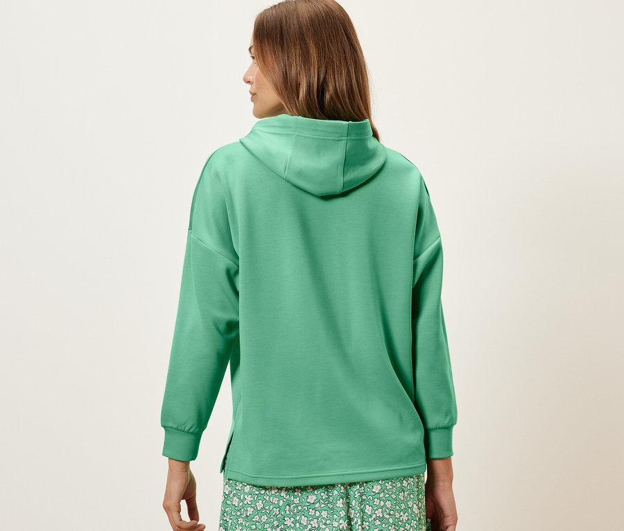 A háttal álló nő zöld, modálkeverékből készült kapucnis felsőt és virágos szoknyát visel.