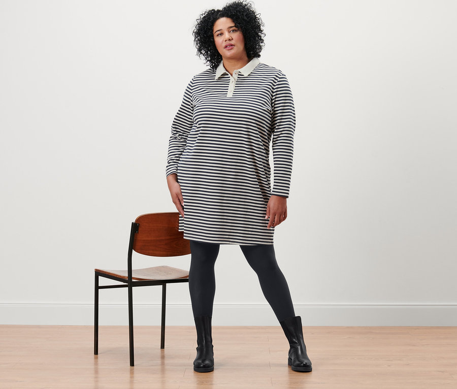 Nő pózol csíkos, póló galléros szabadidőruhában és fekete, 2 darabos, átlátszatlan harisnyanadrágban.