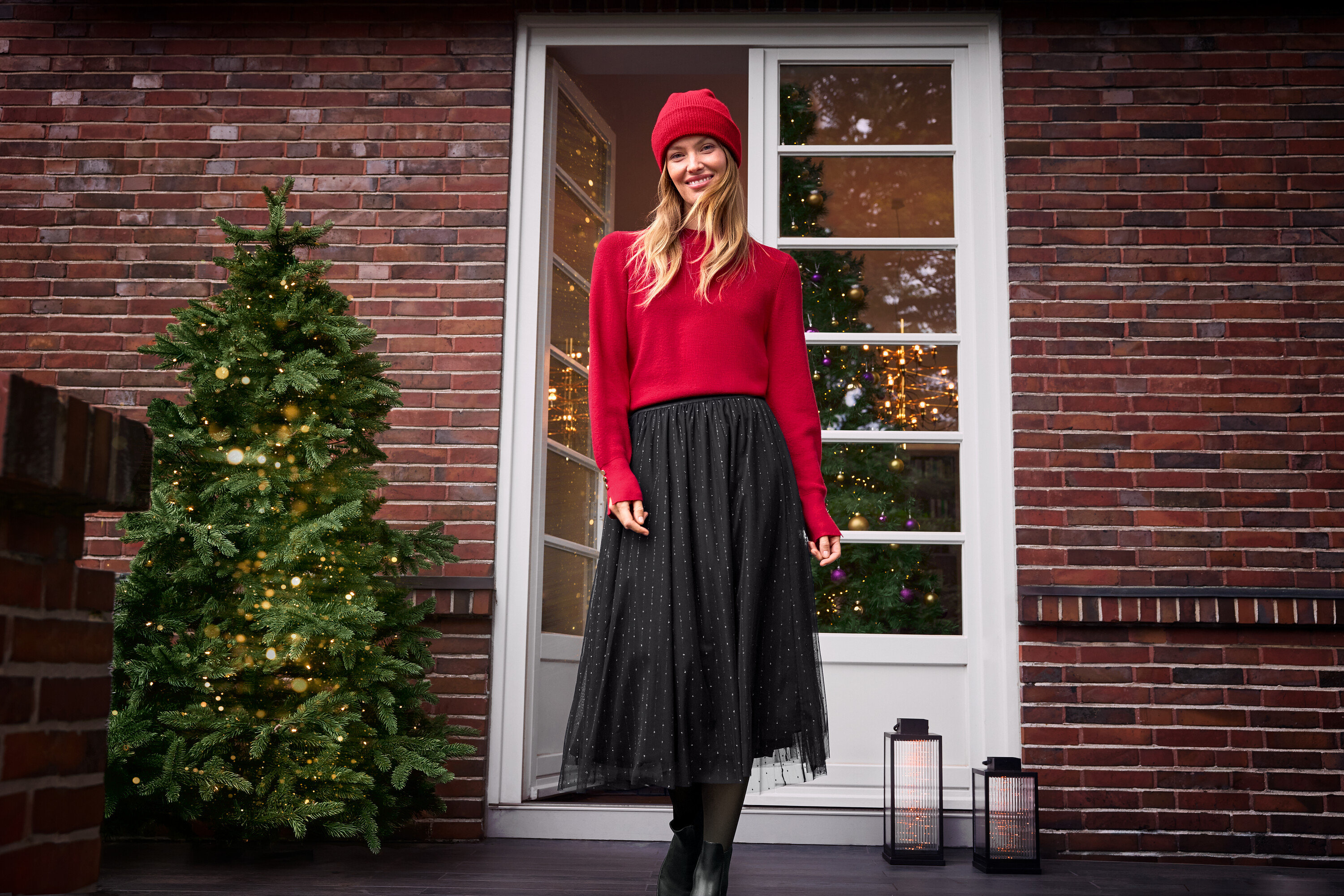 Nő áll a verandán piros pulóverben és fekete tüllszoknyában, bélelt bőrcsizmát és piros sapkát visel.