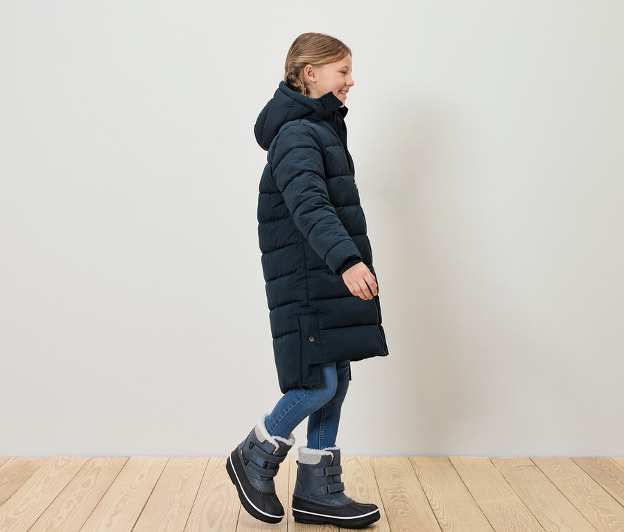 Lány kék steppelt kabátban és hócsizmában sétál a fapadlón.