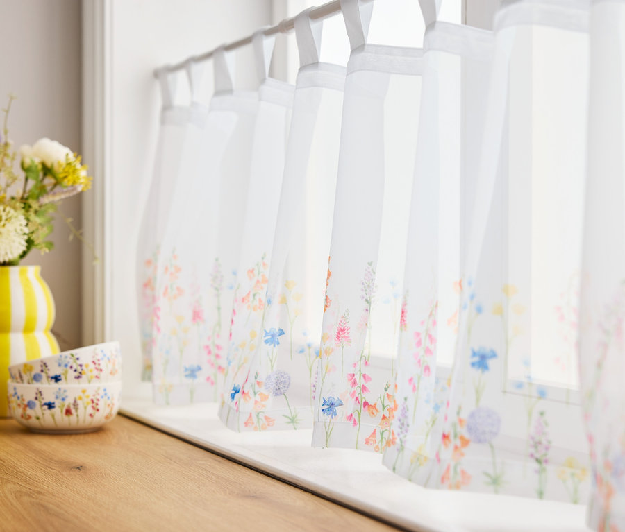 Virágok sárga vázában és két tál fából készült deszkán a virágos függönyös ablakok előtt.