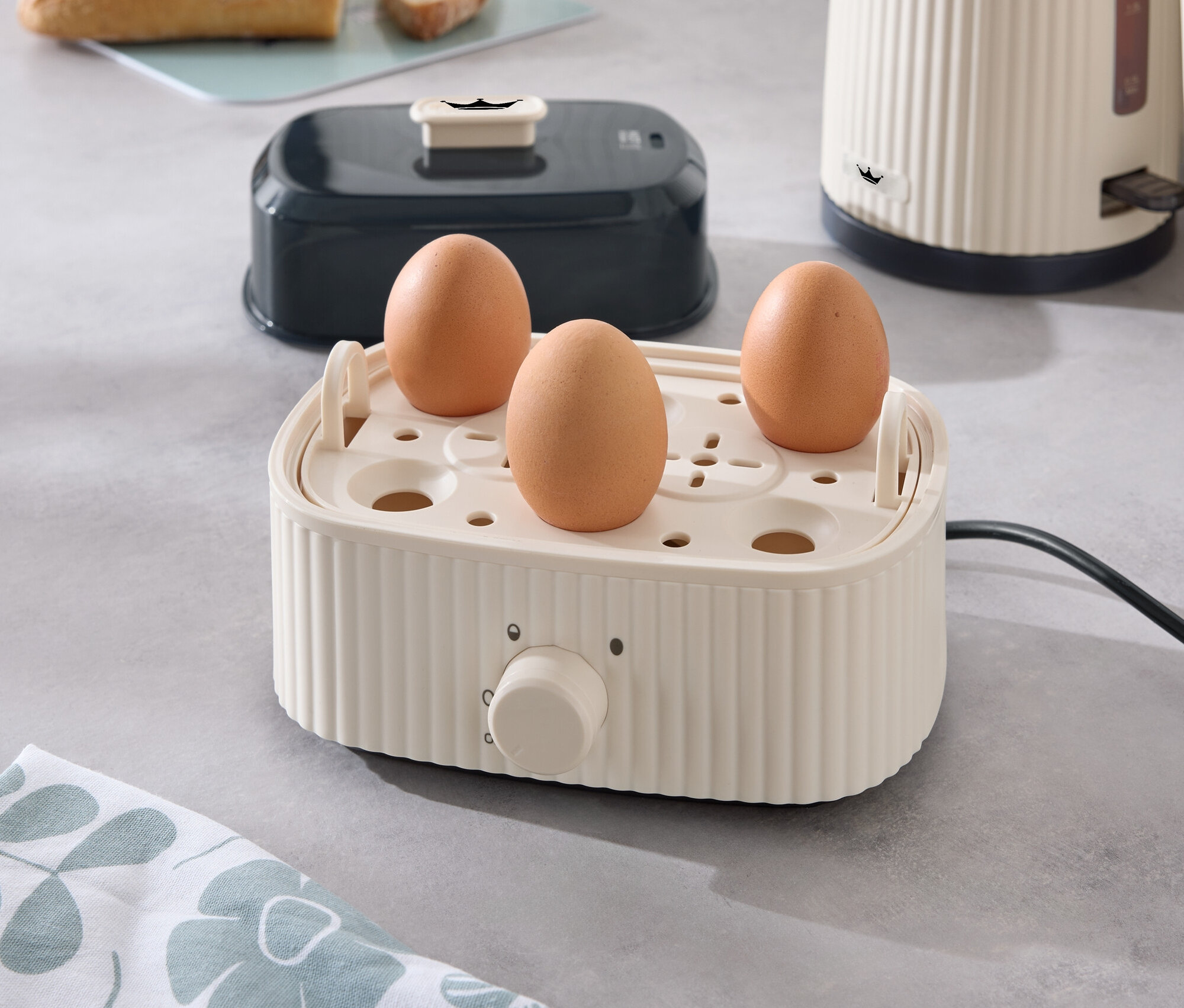 Három tojás egy tojásfőzőben egy konyhapulton, a háttérben egy kenyérpirító, egy vízforraló és kenyér.