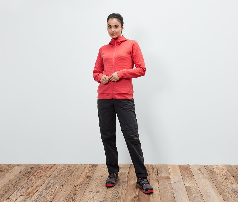 Egy nő korall színű kapucnis felsőben, fekete nadrágban és cipőben pózol egy fapadlón.