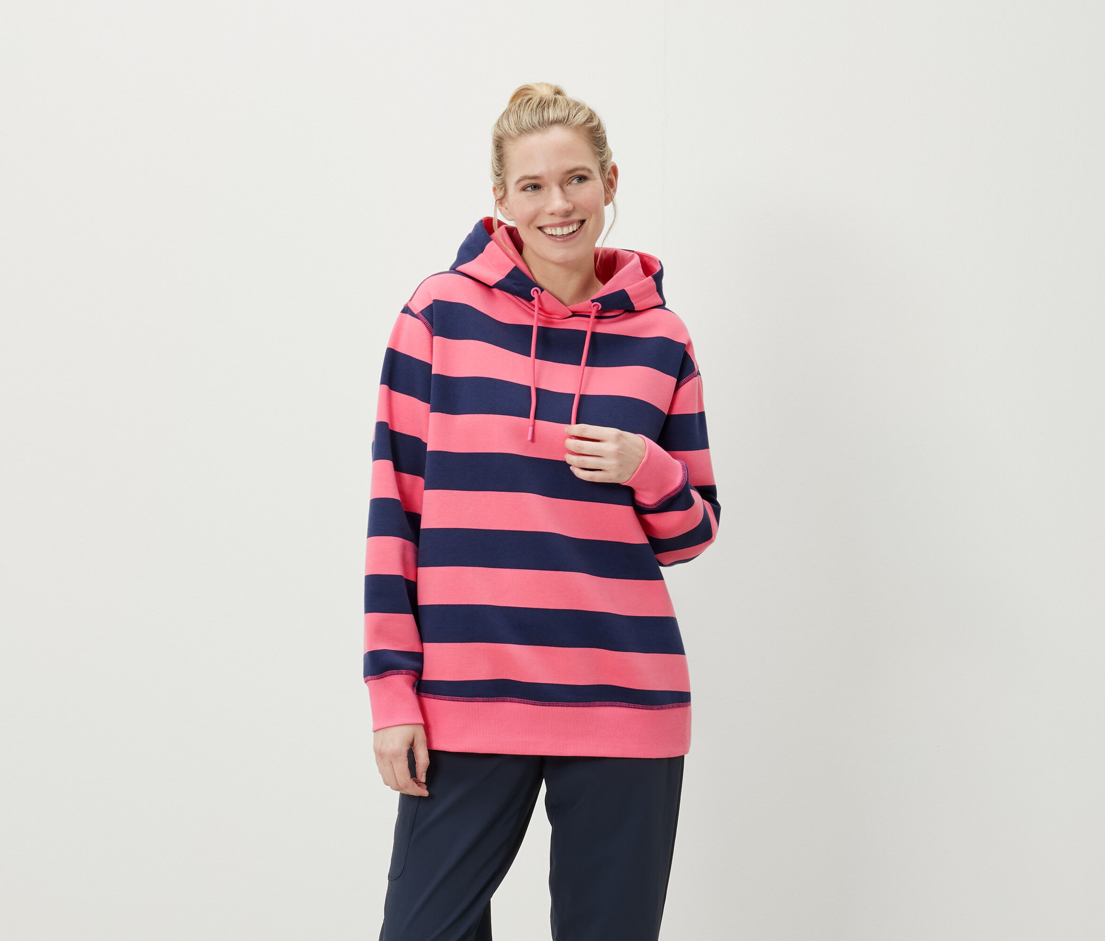 Mosolygó szőke hajú nő rózsaszín-kék csíkos kapucnis pulóvert és sötétkék nadrágot visel.